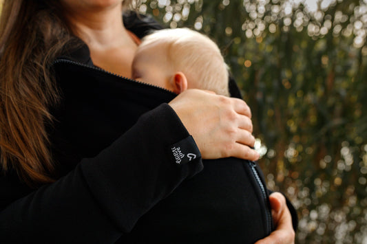 alt="Alvó baba szorosan az anyukájához bújva egy fekete Liliputi 4in1 hordozós pulóverben, közelről látható a Liliputi Love logós mandzsetta